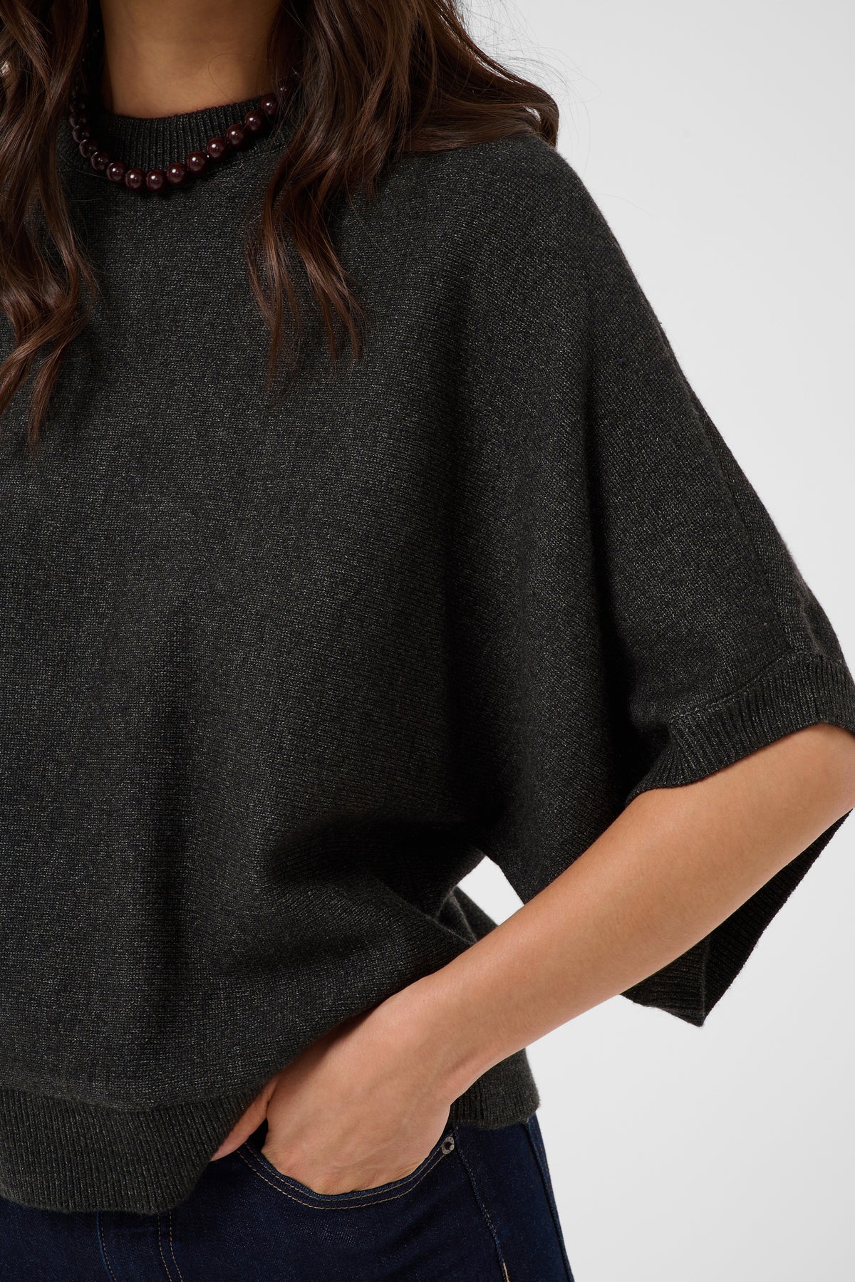 A close-up of a woman wearing a dark charcoal gray knit poncho sweater with a beaded collar, paired with dark blue jeans.