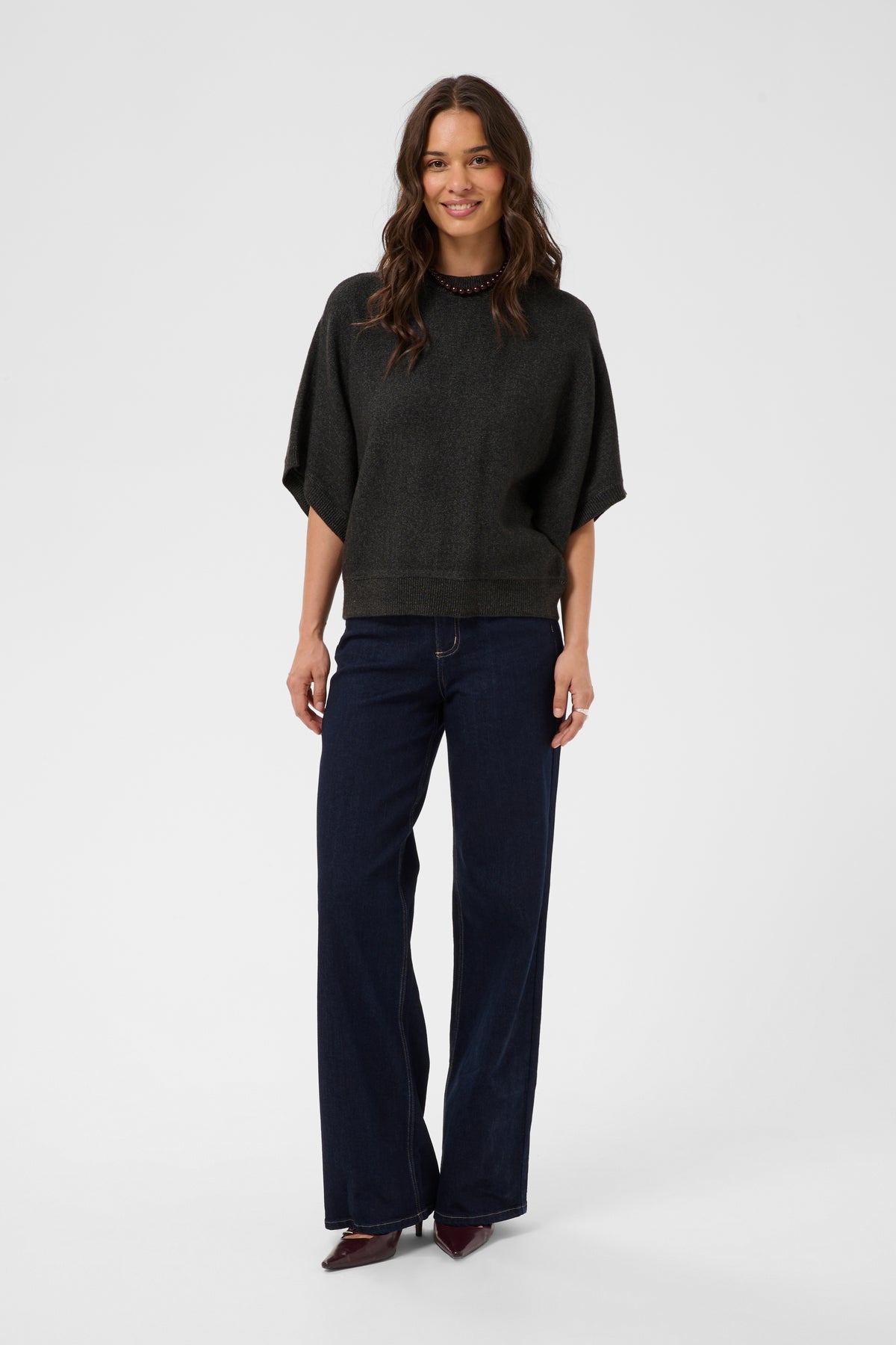 A woman modeling a dark charcoal gray short-sleeve knit top and dark navy wide-leg jeans against a light gray studio background.