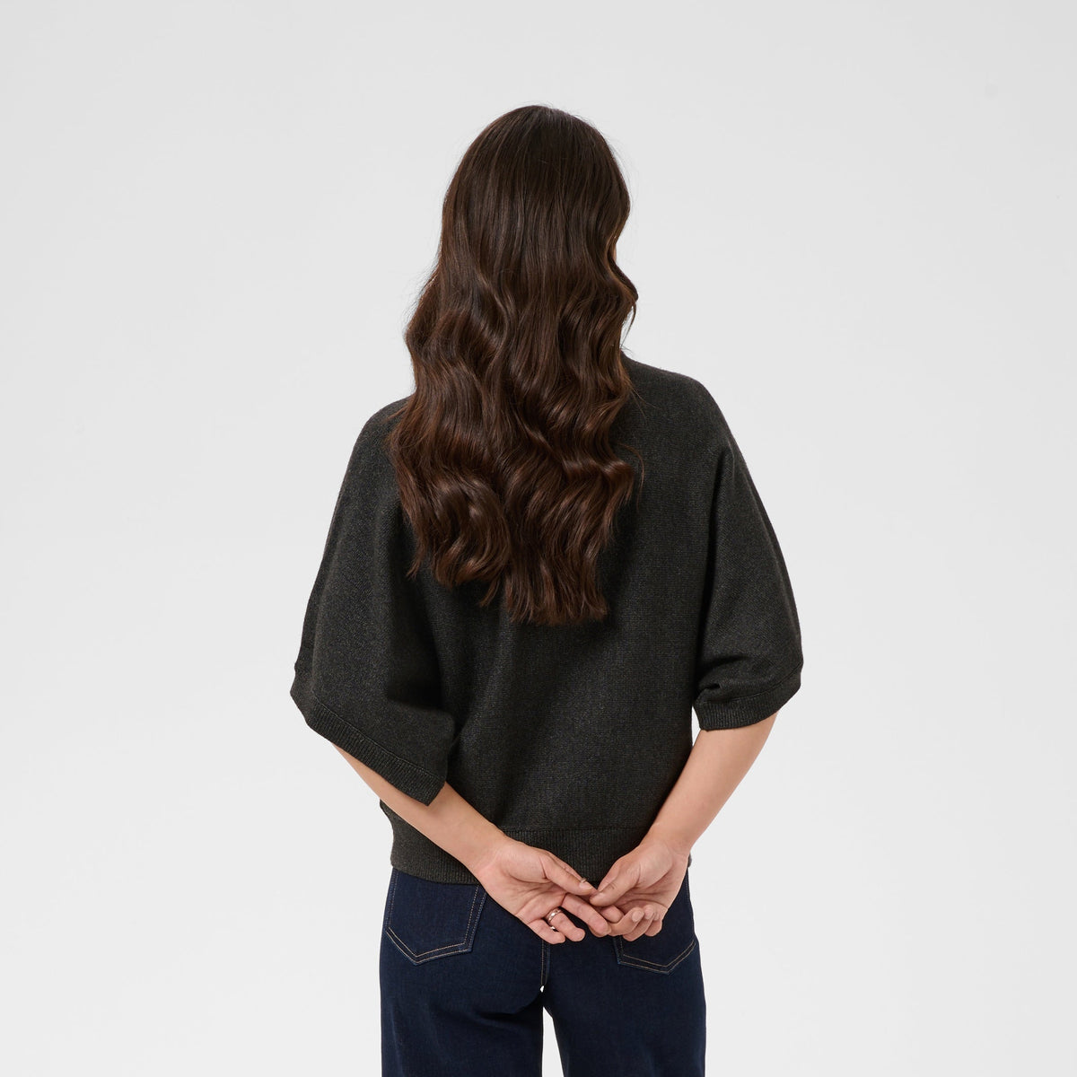 A woman viewed from behind wearing a dark charcoal gray short-sleeve top and dark navy wide-leg jeans, standing against a light gray studio background.