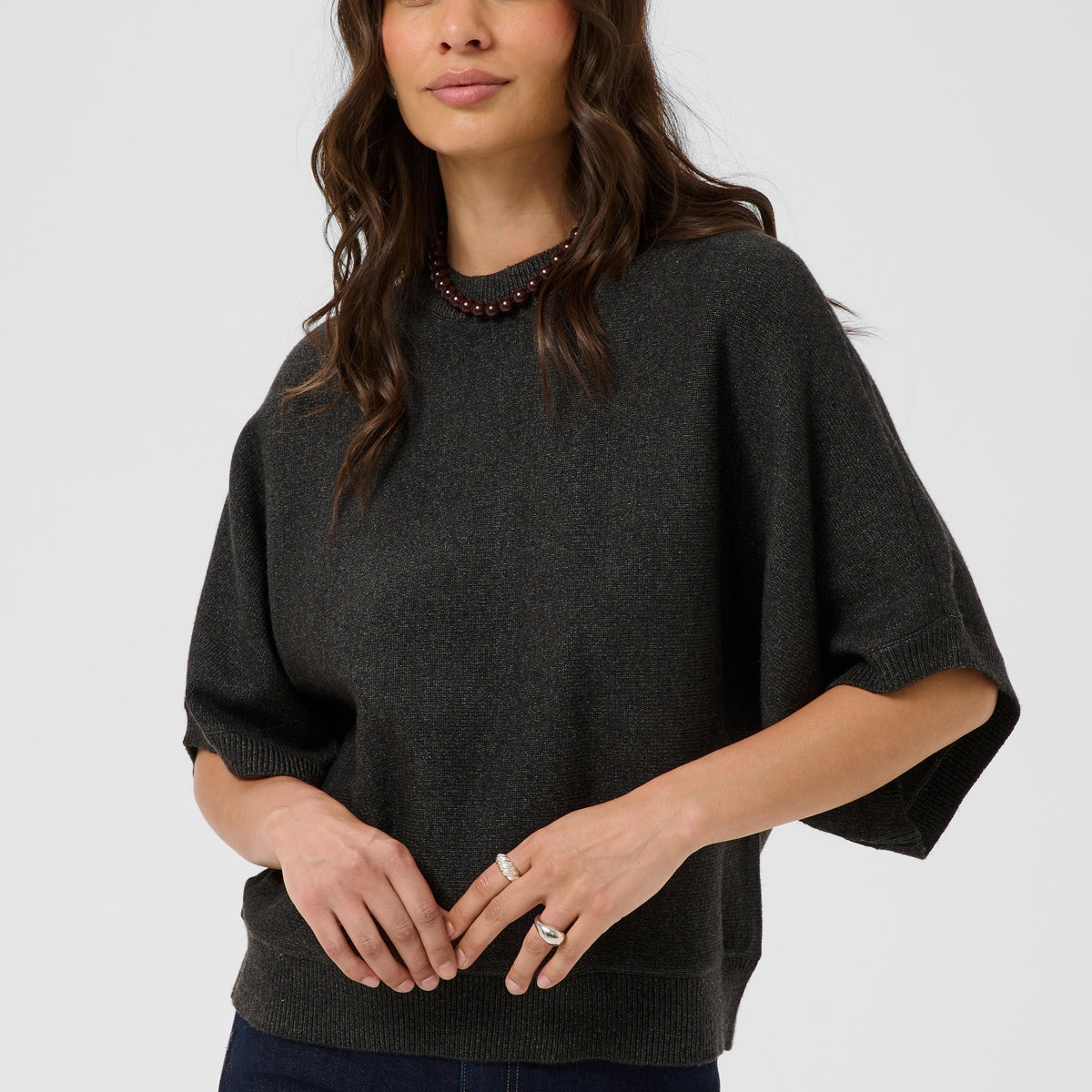 A woman wearing a dark charcoal gray knit sweater with wide, short dolman sleeves and dark blue jeans, standing against a light gray studio background.