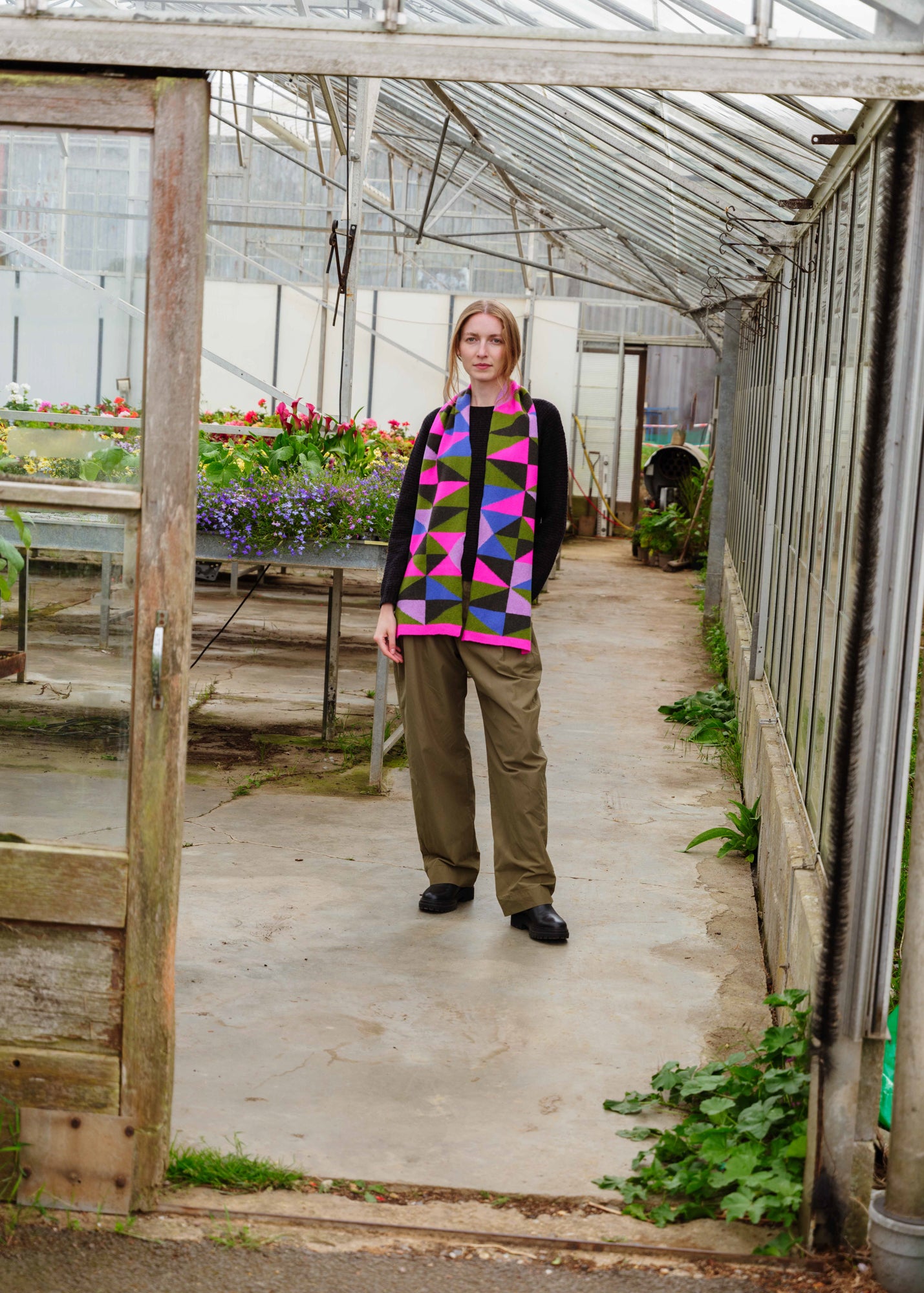 A woman stands in a greenhouse wearing a colorful geometric patterned vest over a black top and olive green trousers.