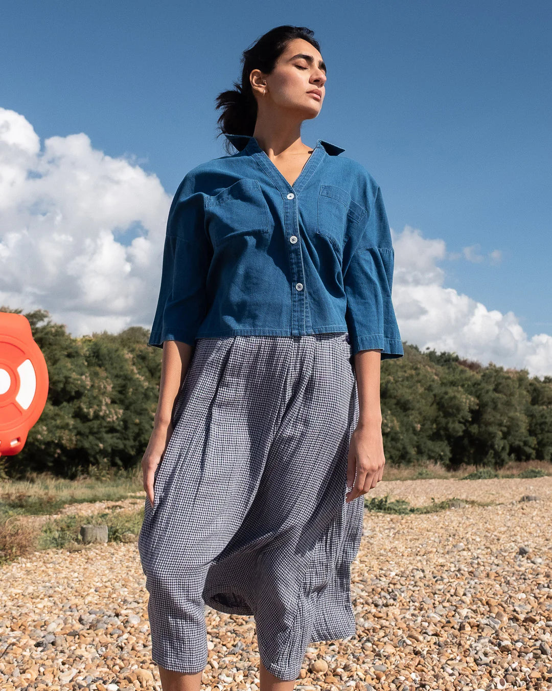 Woman in a blue shirt and gray checkered pants standing outdoors with trees and blue sky in the background