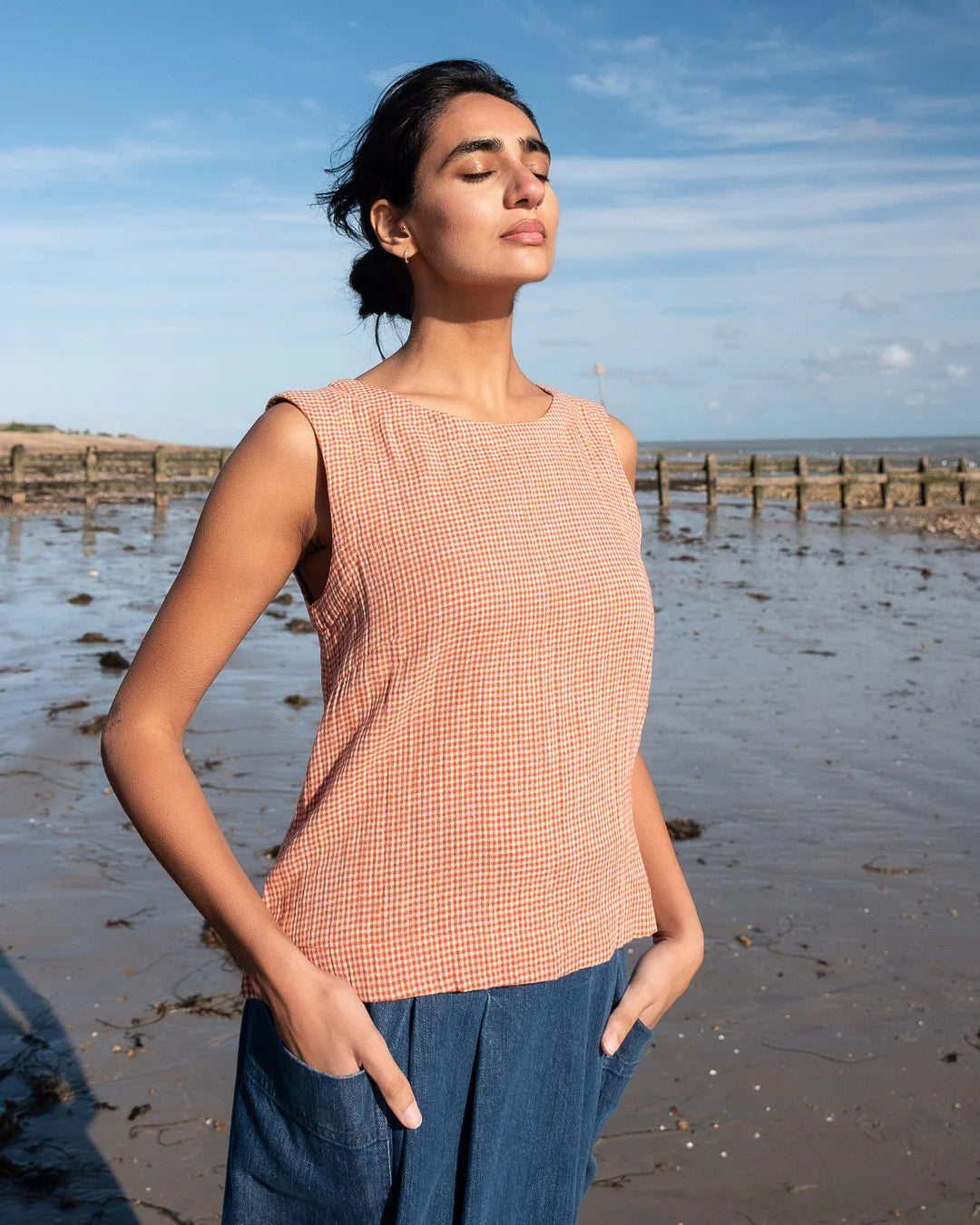 Woman standing on a beach wearing a sleeveless checkered top and blue pants.