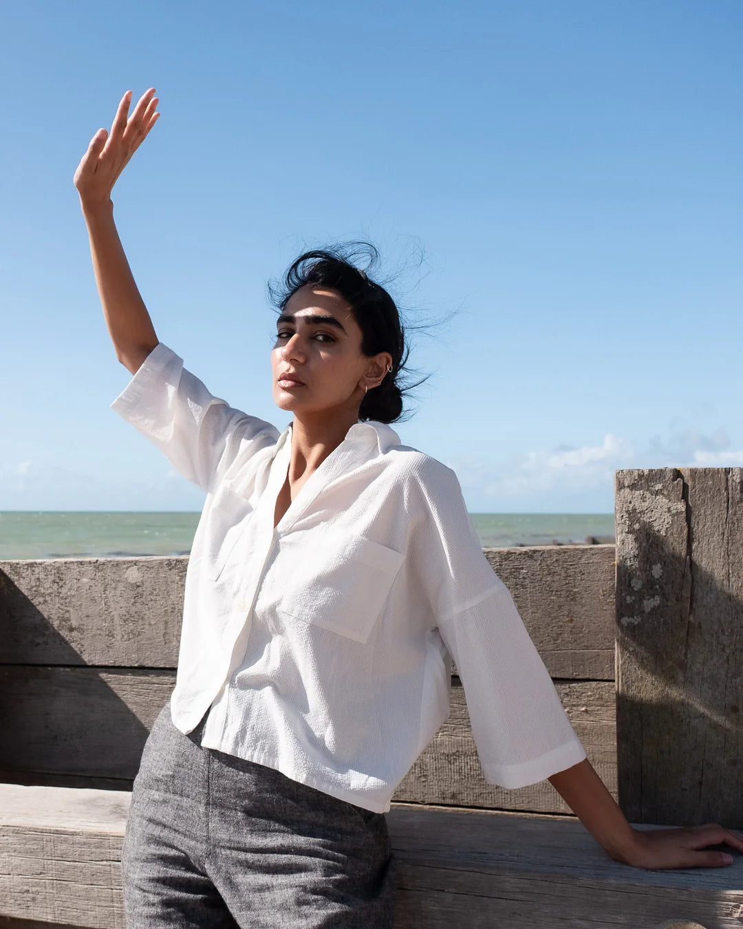 Woman in a white shirt and gray pants standing on a wooden platform with a clear blue sky and ocean in the background.