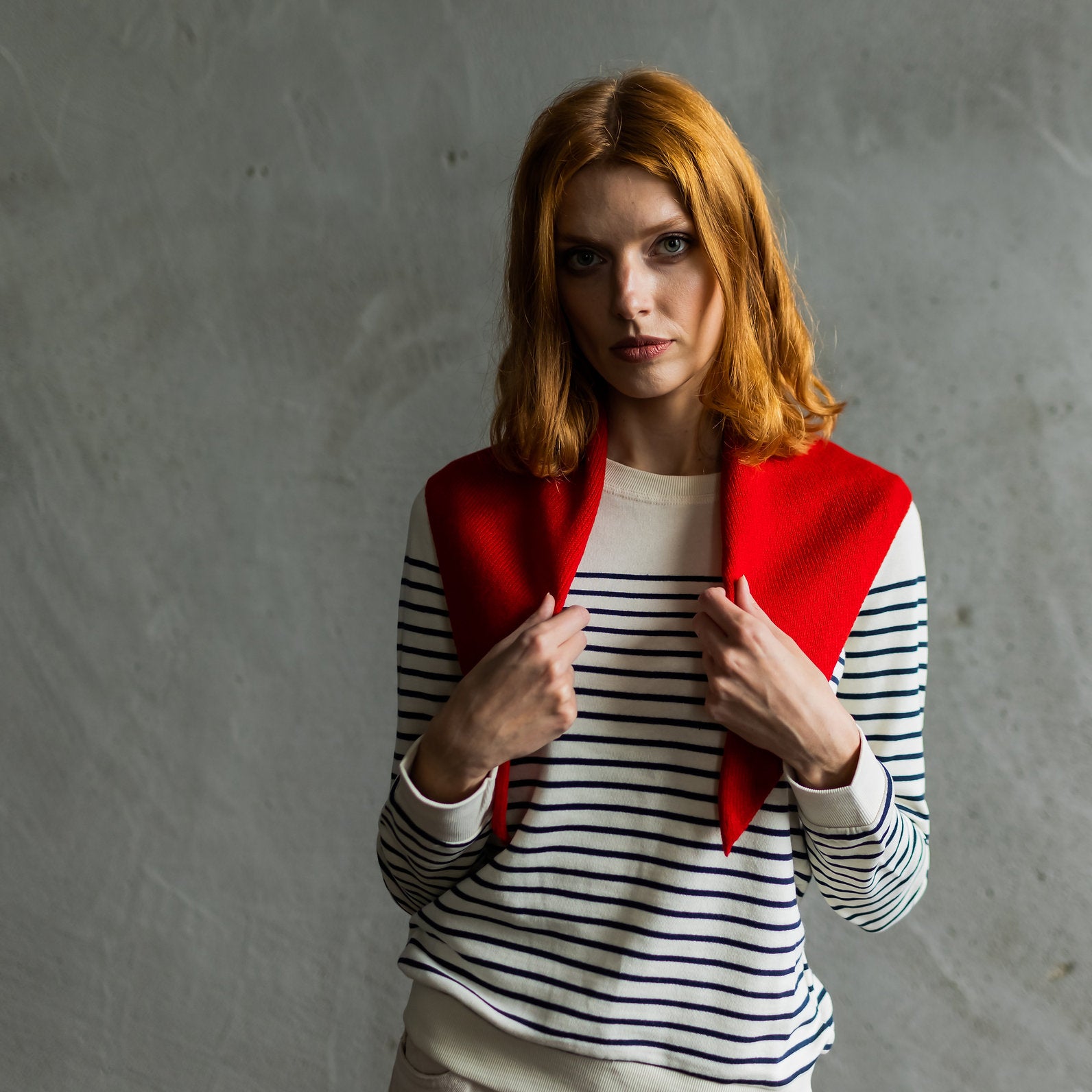 A woman with red hair wearing a red knit cardigan over a black and white striped long-sleeve top, posing against a textured gray concrete wall.