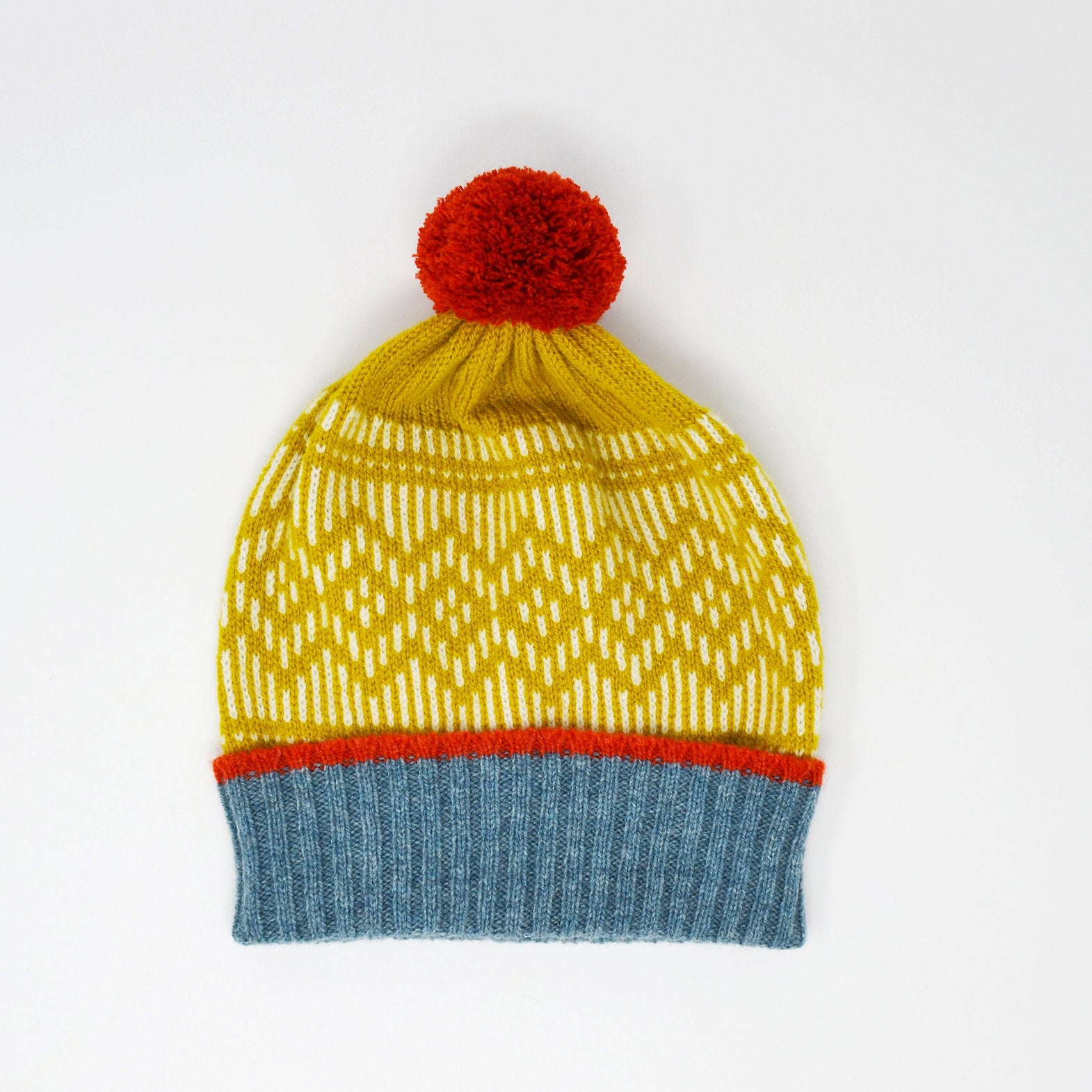 Mustard yellow knitted beanie with white and orange detailing, featuring a red pompom on top and a blue ribbed cuff.