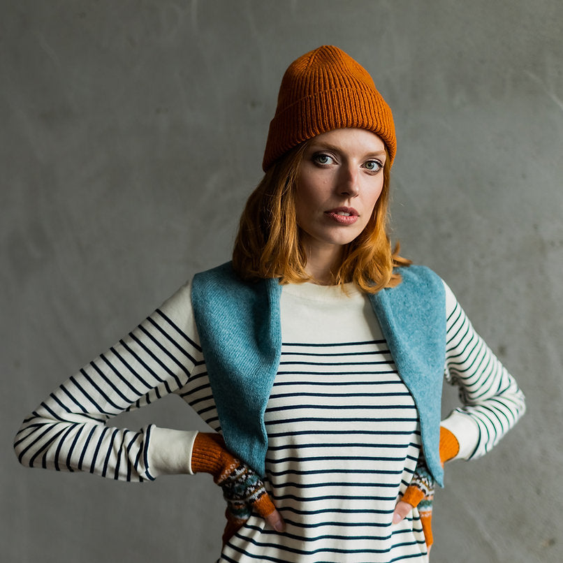 Woman wearing a ribbed orange beanie, striped cream and navy sweater, teal shawl, and orange fingerless gloves.