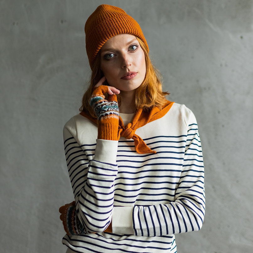 Woman wearing an orange knit beanie and patterned fingerless gloves with a cream and navy striped sweater.