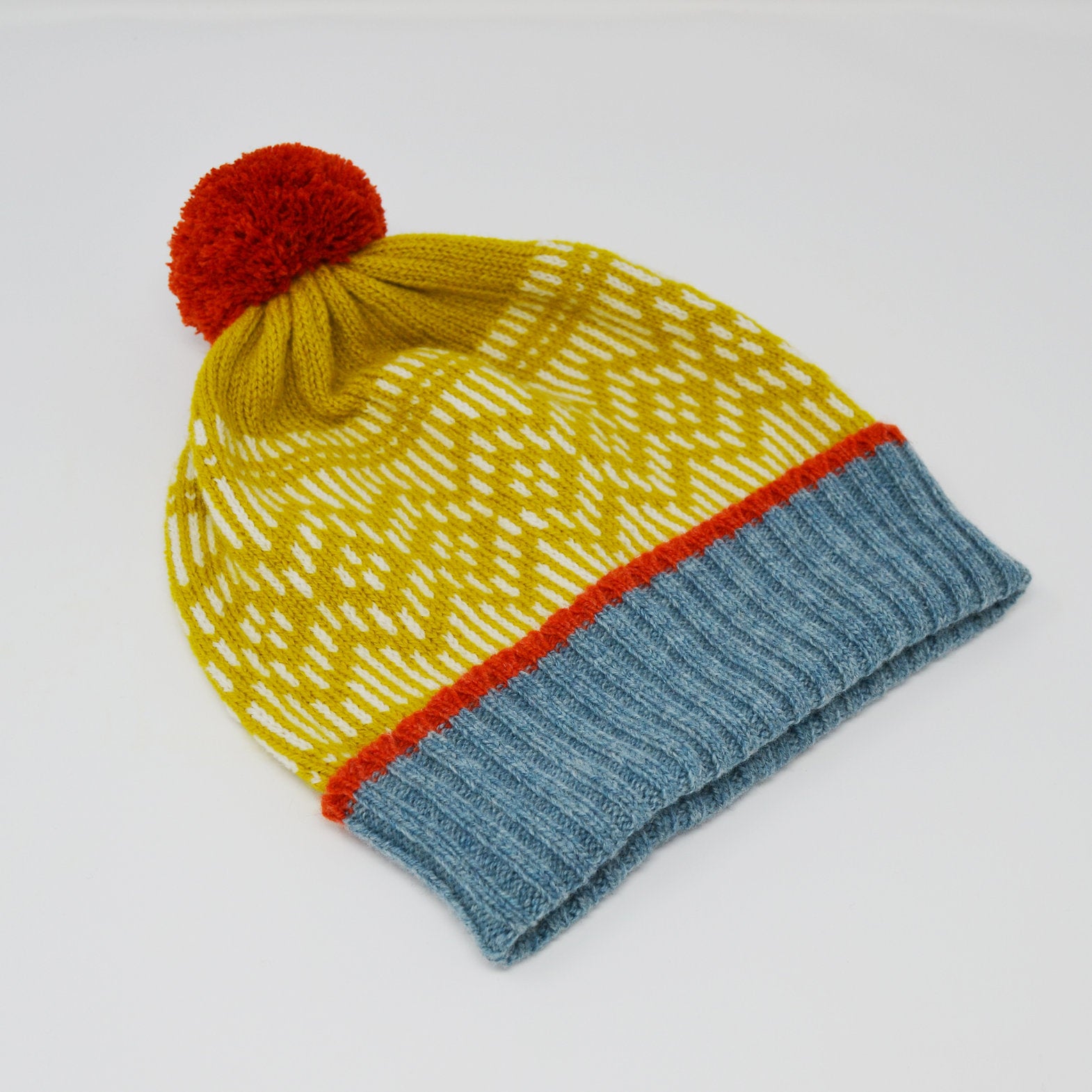 Knit beanie with a mustard yellow body, white pattern, orange and blue trim, and a red pompom.