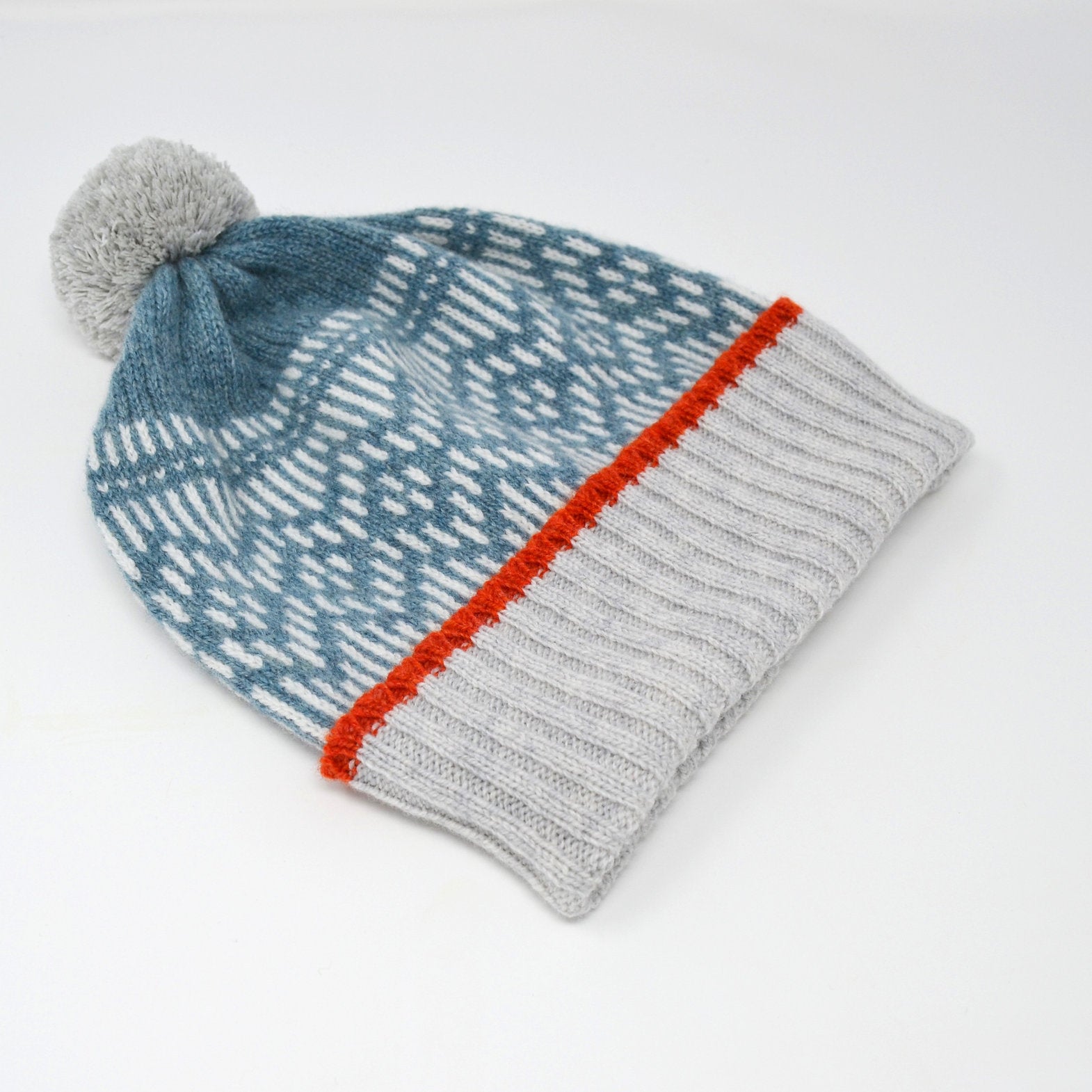 Knit beanie with grey pompom, teal and white patterned crown, and grey ribbed cuff accented with orange stitching.