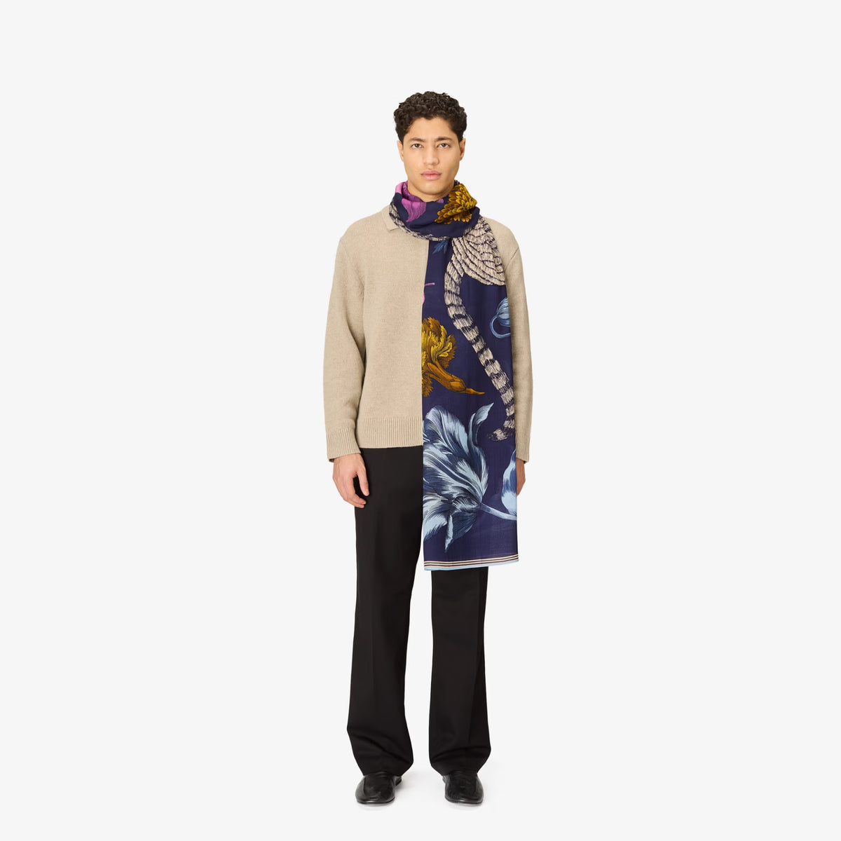 A male model stands against a plain light gray studio background wearing a beige cardigan, black trousers, and a long printed scarf draped over his left shoulder. The scarf has a dark navy base with large white floral shapes and mustard-yellow and purple accents.