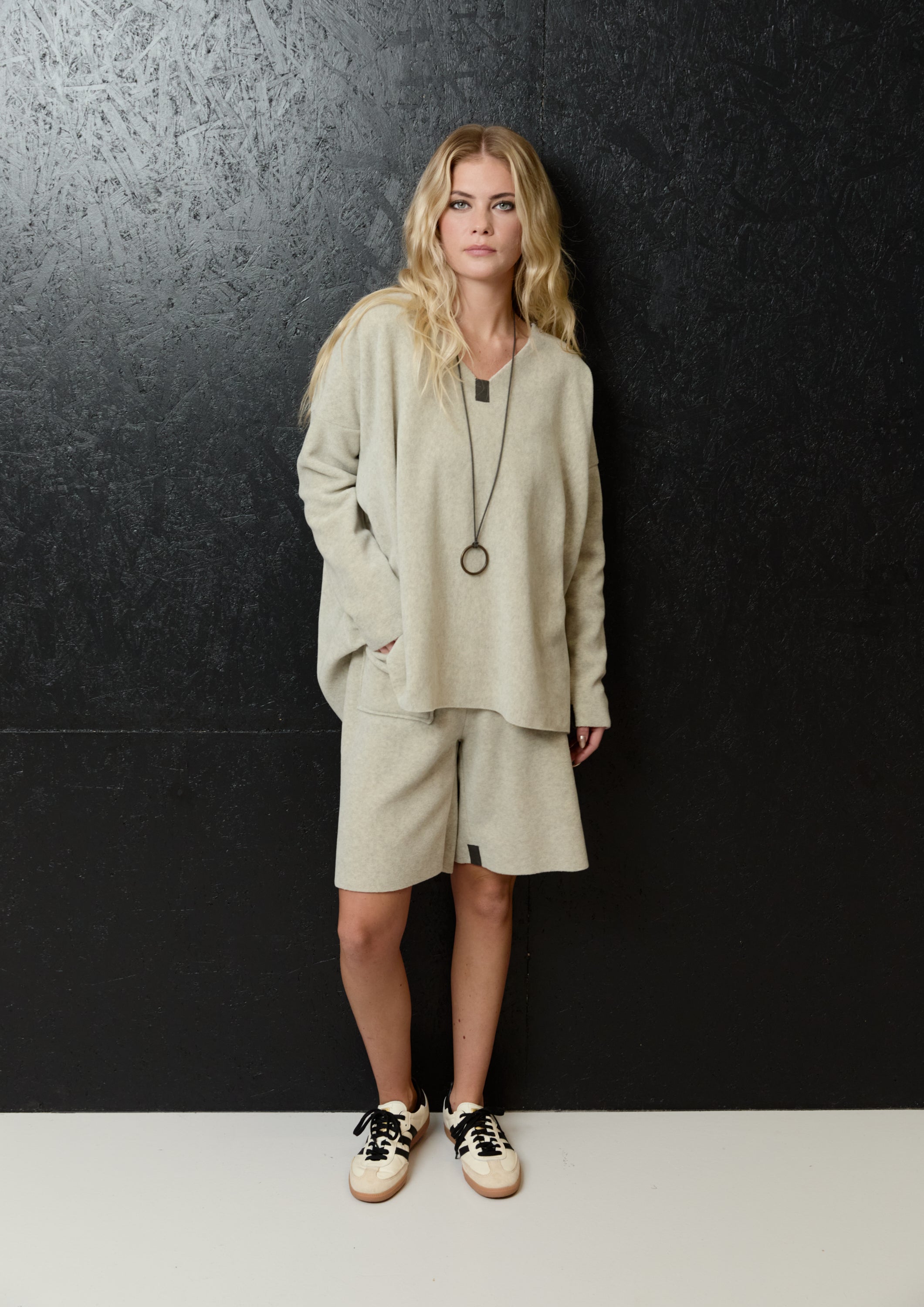 A woman stands against a dark textured wall wearing a light beige knit lounge set consisting of a long-sleeve top and matching shorts, accessorized with a long pendant necklace and black-and-white sneakers.