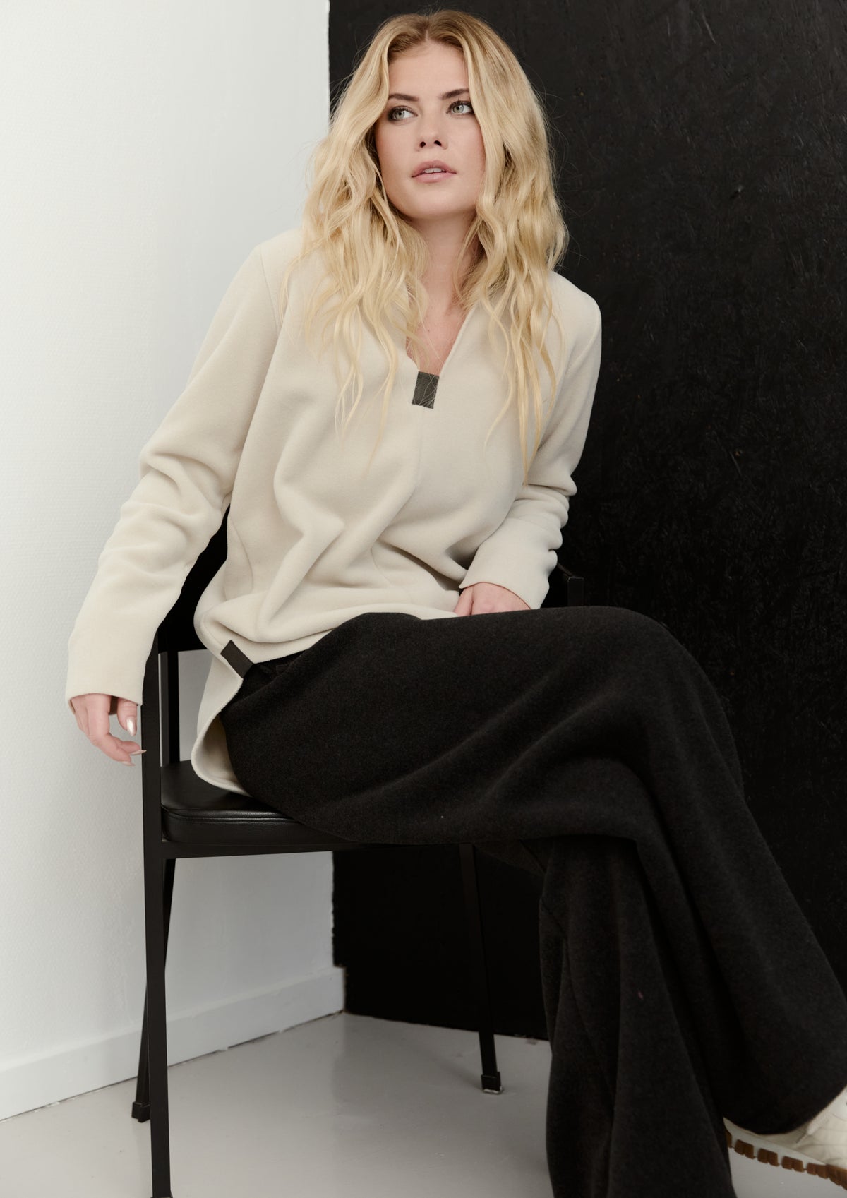 Woman sitting on a chair wearing a beige sweater and black pants against a white wall.