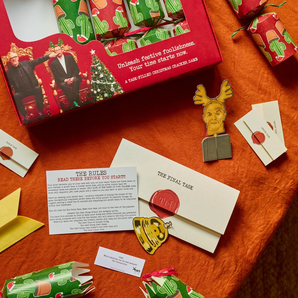 A Christmas-themed game set laid out on an orange fabric background, including a red box with festive illustrations, wrapped gift boxes, cards, envelopes with red wax seals, and small decorative pieces.