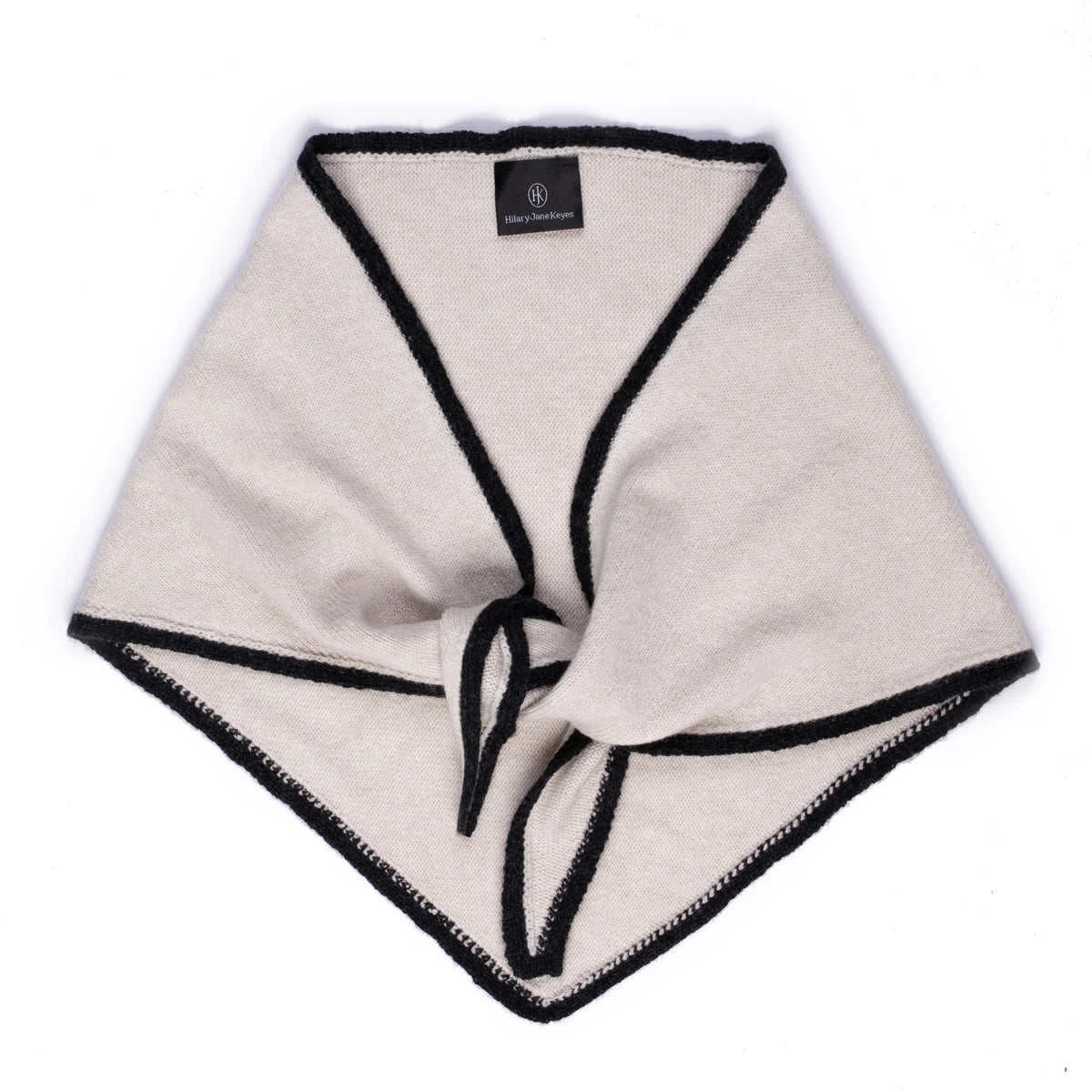 A light beige knit wrap or poncho with black trim and a front tie detail, laid flat on a white background. A small black brand label is visible inside the neckline.