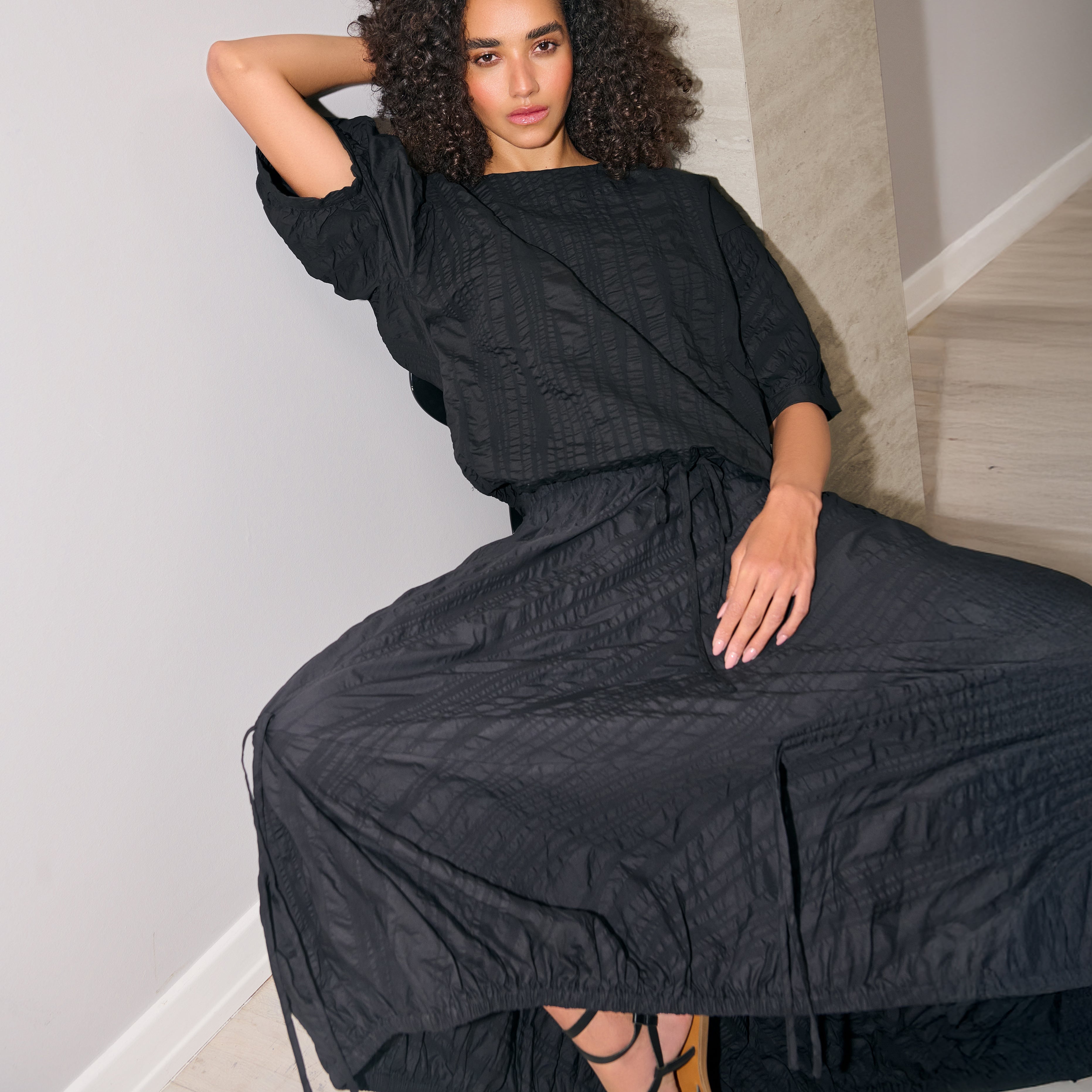 Woman wearing a black textured dress sitting on a white wall.