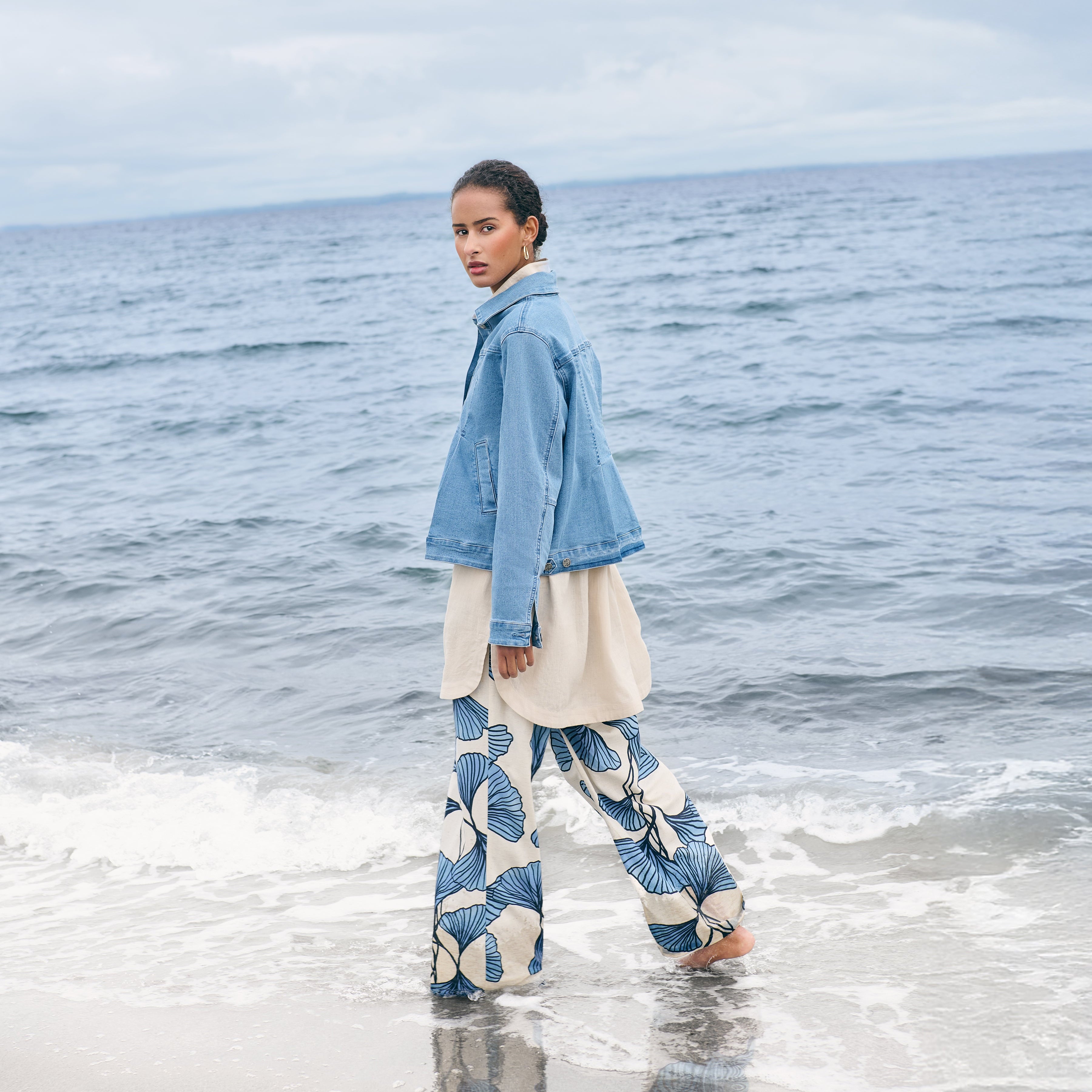 Person wearing a blue jacket and patterned pants standing on a beach with ocean in the background