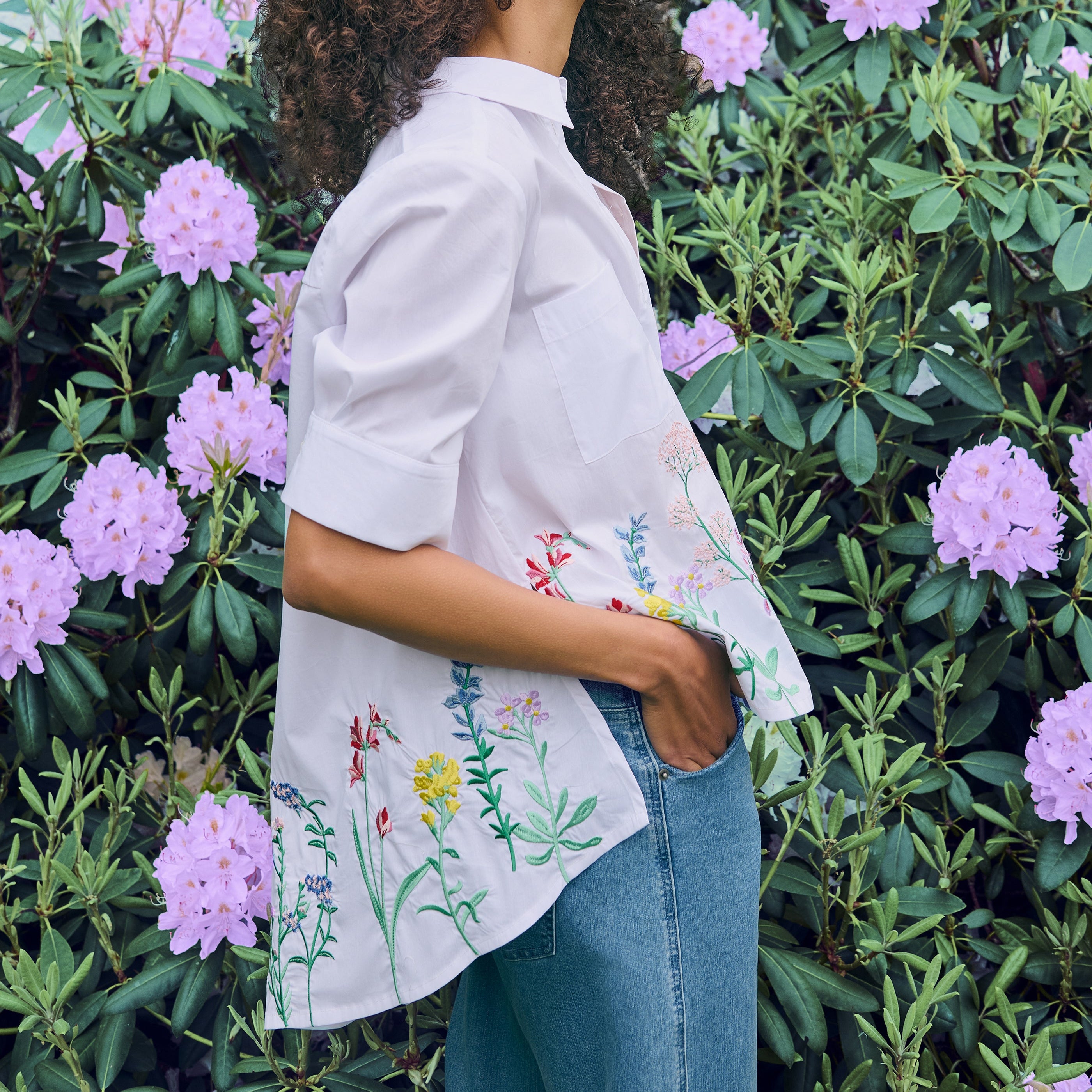 Woman wearing a white blouse with floral embroidery standing in front of pink flowers.