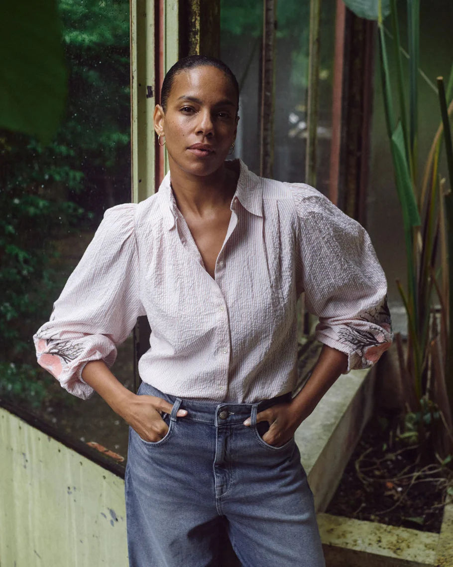 Person wearing a white textured shirt and blue jeans standing outdoors with greenery in the background