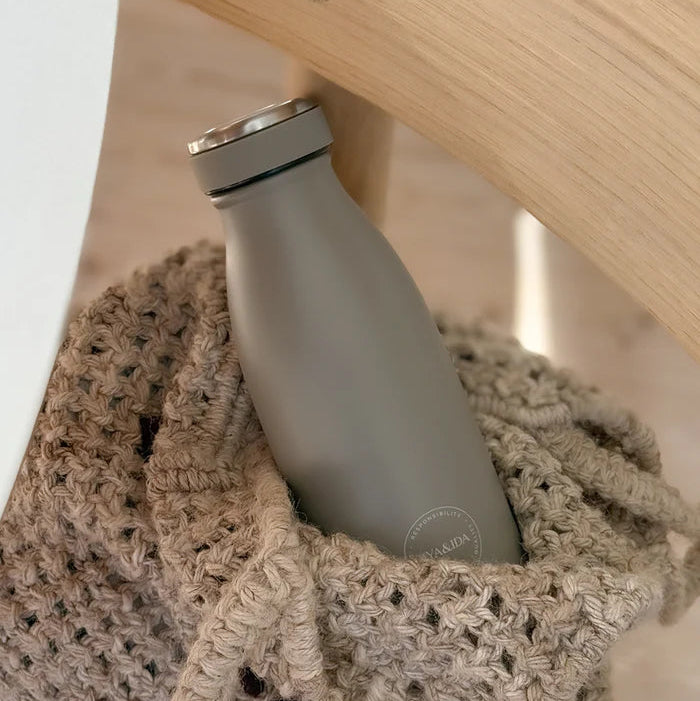 Gray water bottle with a silver cap inside a textured beige bag on a wooden surface.