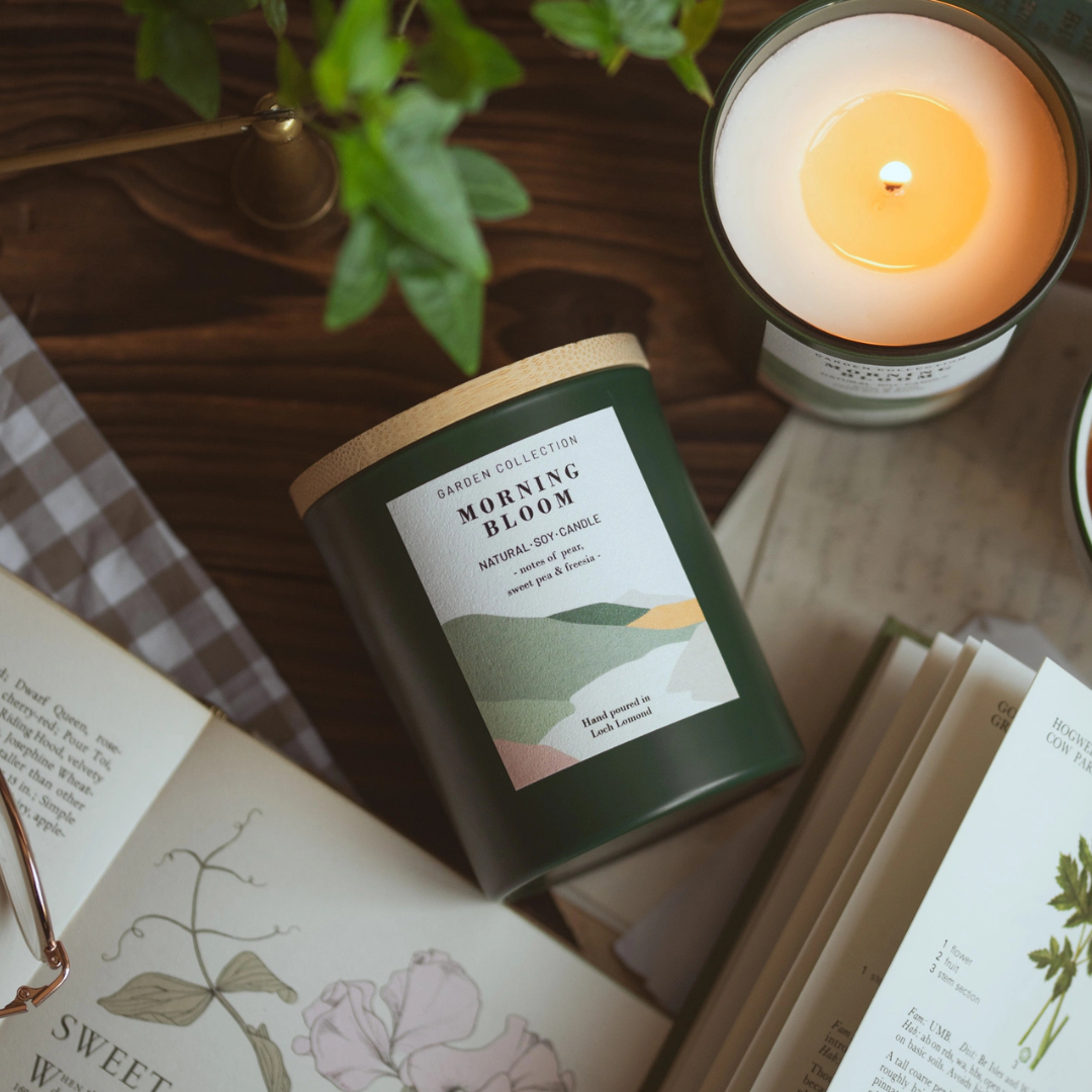 A dark green candle jar labeled 'GARDEN COLLECTION MORNING BLOOM' sits on a wooden table surrounded by books, a lit candle, and green leaves in a styled flat-lay setting.