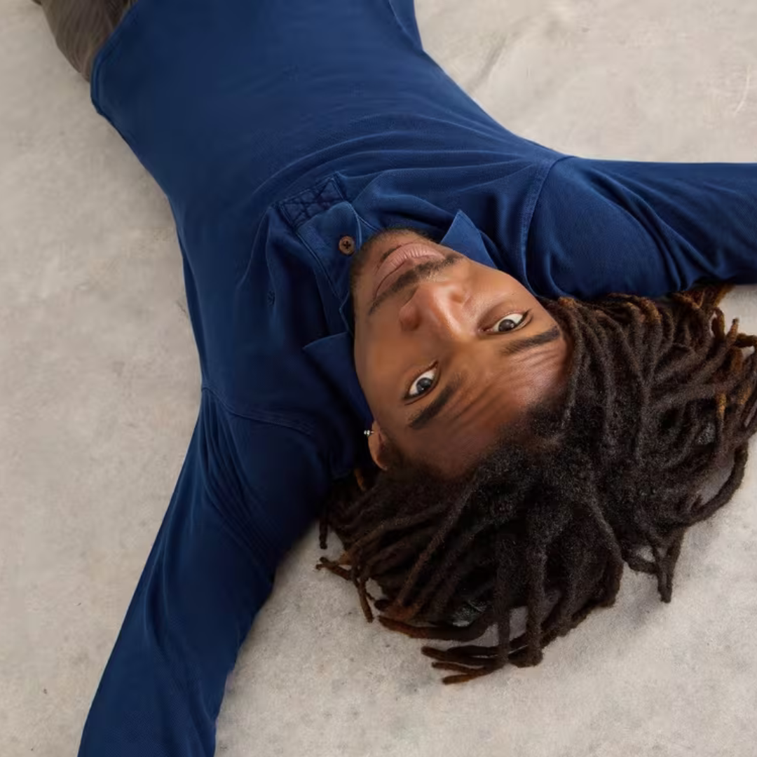 A person with long dreadlocks lies on their back wearing a dark blue long-sleeved polo shirt.