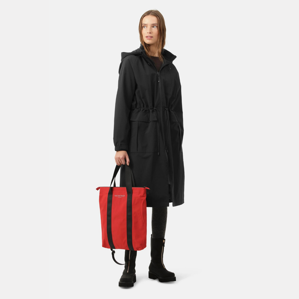 Woman in black hooded parka holding a red Rains tote bag. The bag features black straps and subtle branding. She wears dark