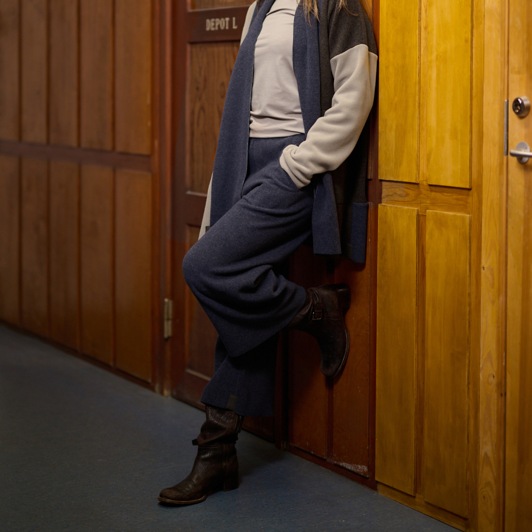 Person leaning against a wooden door wearing a blue vest and gray pants.