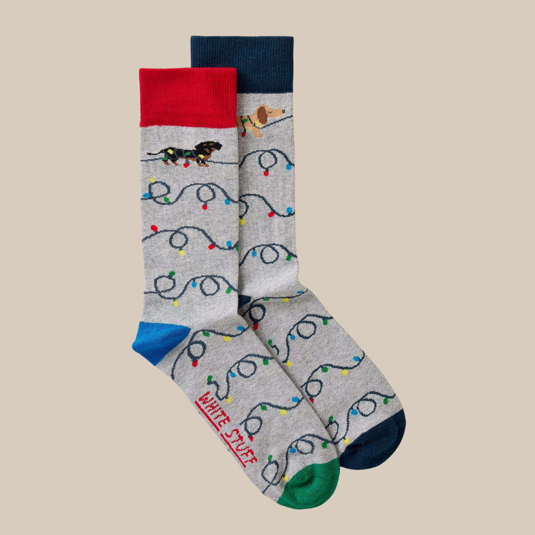 A pair of gray crew socks with a holiday Christmas lights and dachshund pattern, featuring a red cuff, blue heel, green toe, and the words 'WHITE STUFF' printed on the sole.