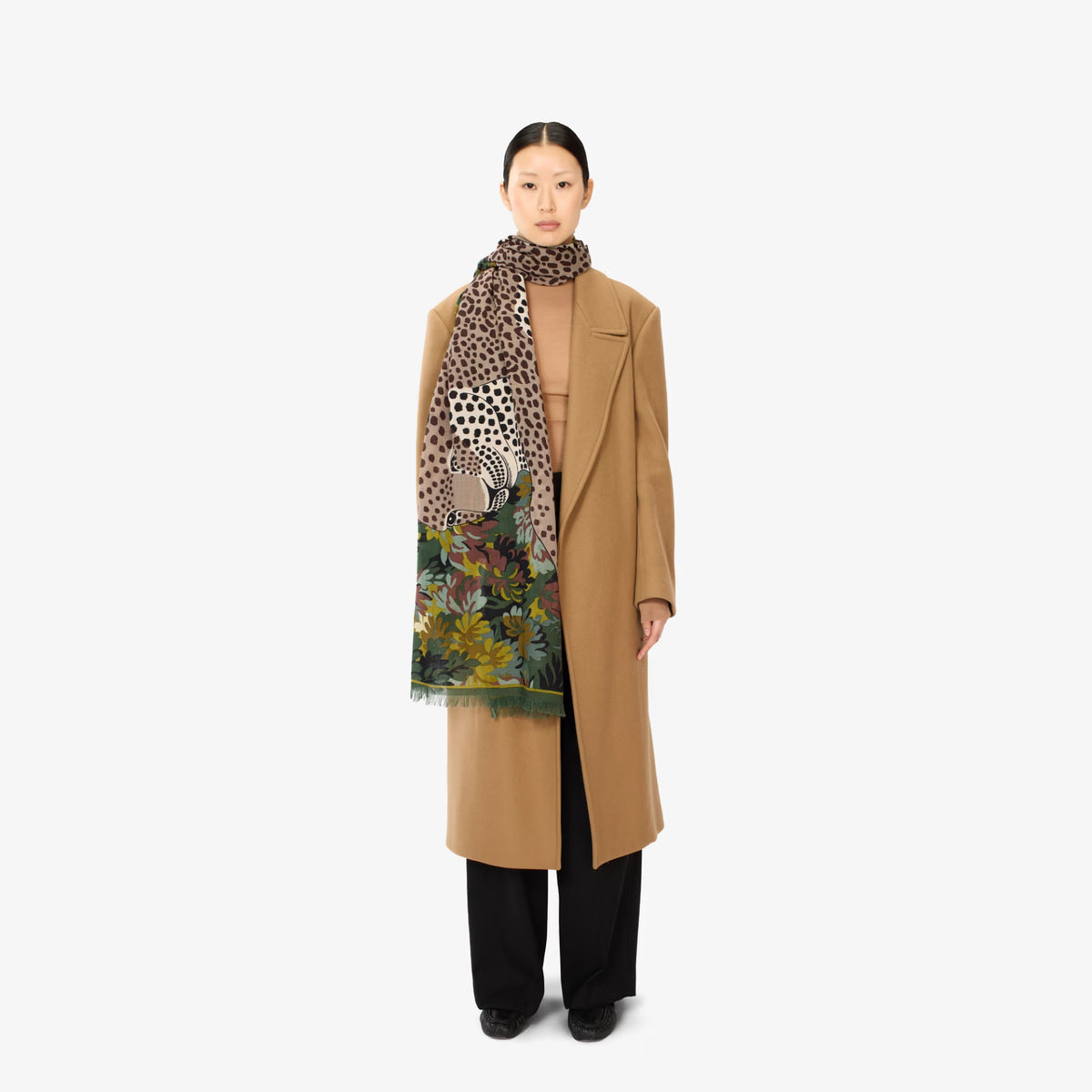 A model stands against a light gray background wearing a long camel coat, black wide-leg trousers, and a patterned scarf featuring a leopard print and a colorful floral/garden illustration.