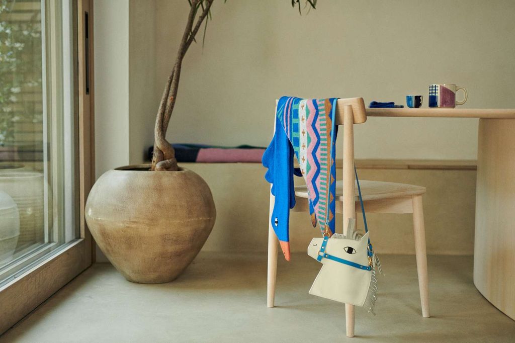 A styled interior scene with a light wood dining table, chair, and large ceramic planter. A colorful patterned scarf and a blue-and-white plush toy are draped over the chair, alongside a small white horse-shaped handbag with blue trim.