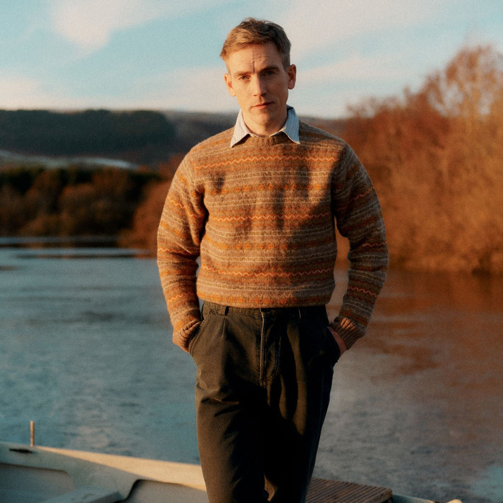 a male model with hands in pockets stood by the side of a loch wearing a Harley of Scotland Men's Jumper - Nutmeg