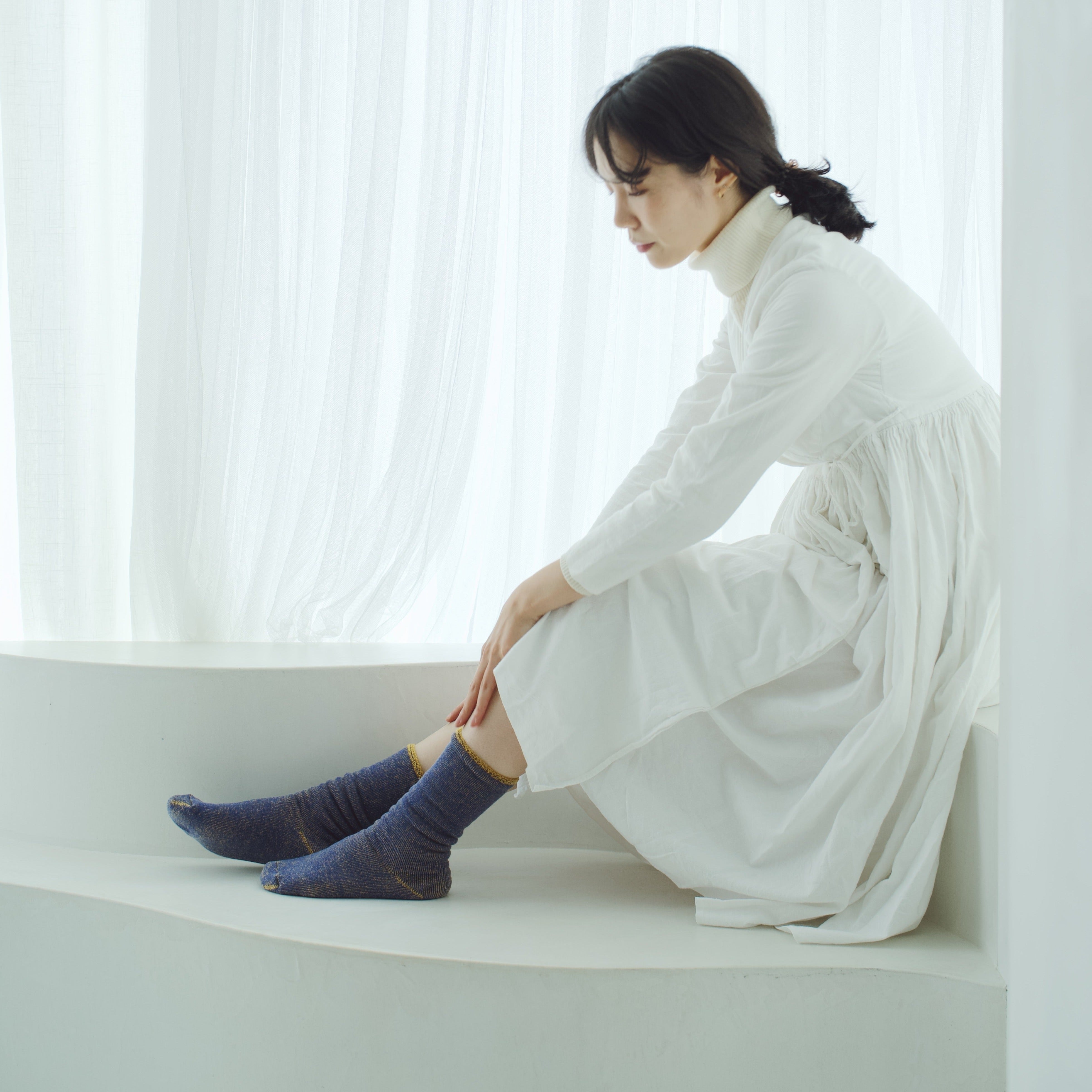 Woman in a white dress and blue socks sits on a white window seat.
