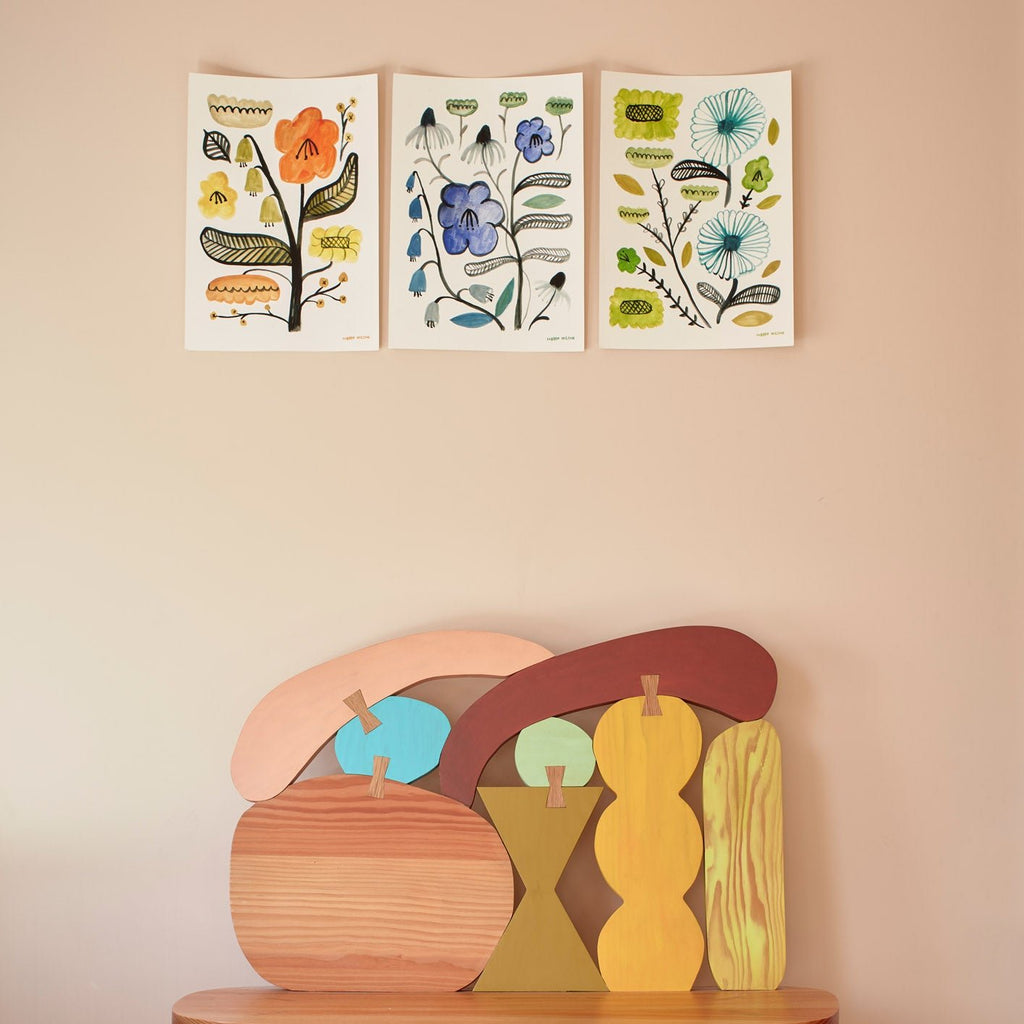 Three botanical prints by Donna Wilson feature orange, blue, and green designs on a beige wall above colorful wooden shapes.