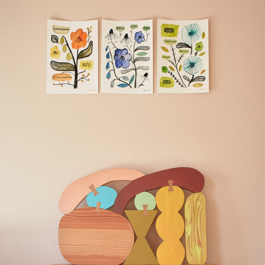 Three floral art prints by Donna Wilson in yellow, blue, and green, with colorful wooden shapes below.