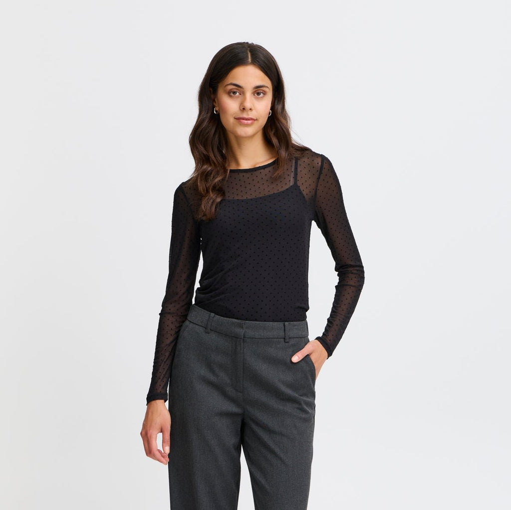 A woman wears a black dotted long-sleeve top by Fransa and gray tailored trousers, posing confidently.
