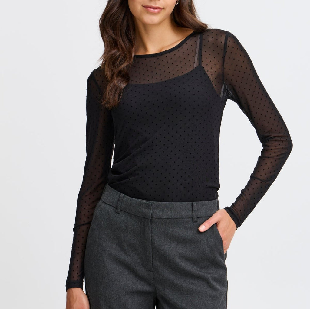 A woman in a black dotted sheer top by Fransa, paired with grey trousers, poses confidently.