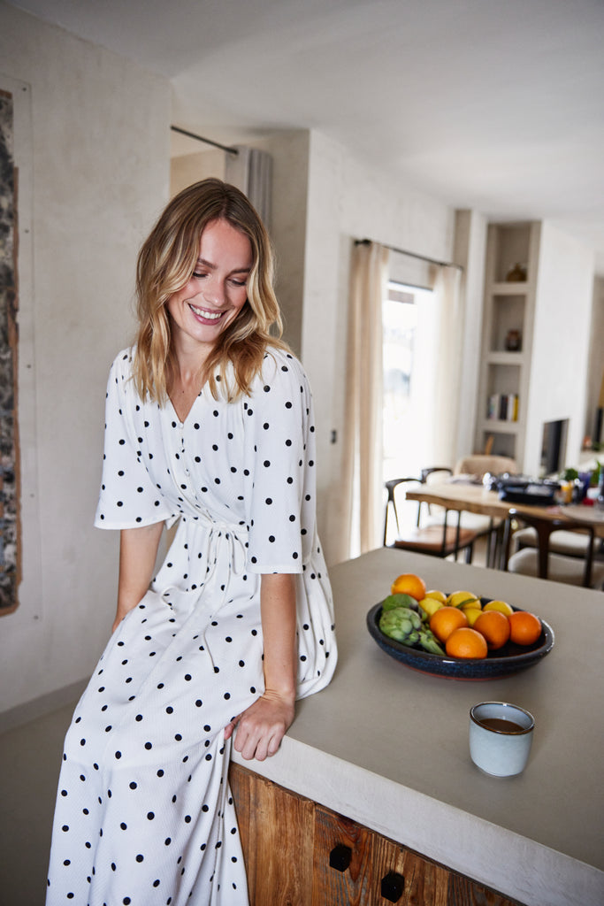 A woman smiles in a white polka dot dress by Fransa, seated on a kitchen counter beside a bowl of fruit.