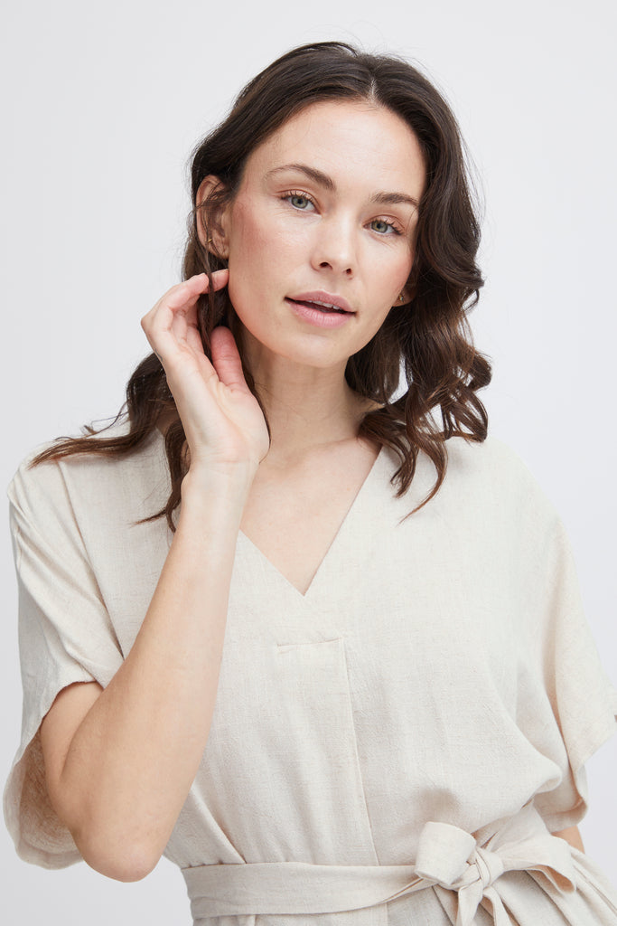 A woman in a light beige Fransa dress with a v-neck and tied waist, gently touching her hair.