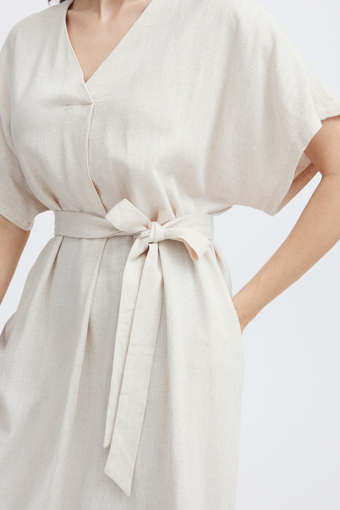 A woman wears a light beige Fransa dress with a v-neck and a tied waist, showcasing a relaxed, elegant style.