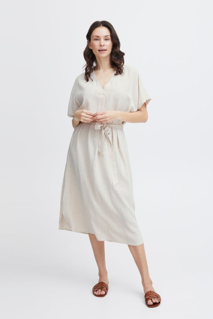 A woman in a light beige Fransa dress, featuring a belt, with brown sandals, standing against a white background.