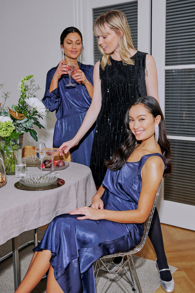 Three women celebrate together in Fransa outfits. One wears a navy satin dress; another dons a black sequined dress.