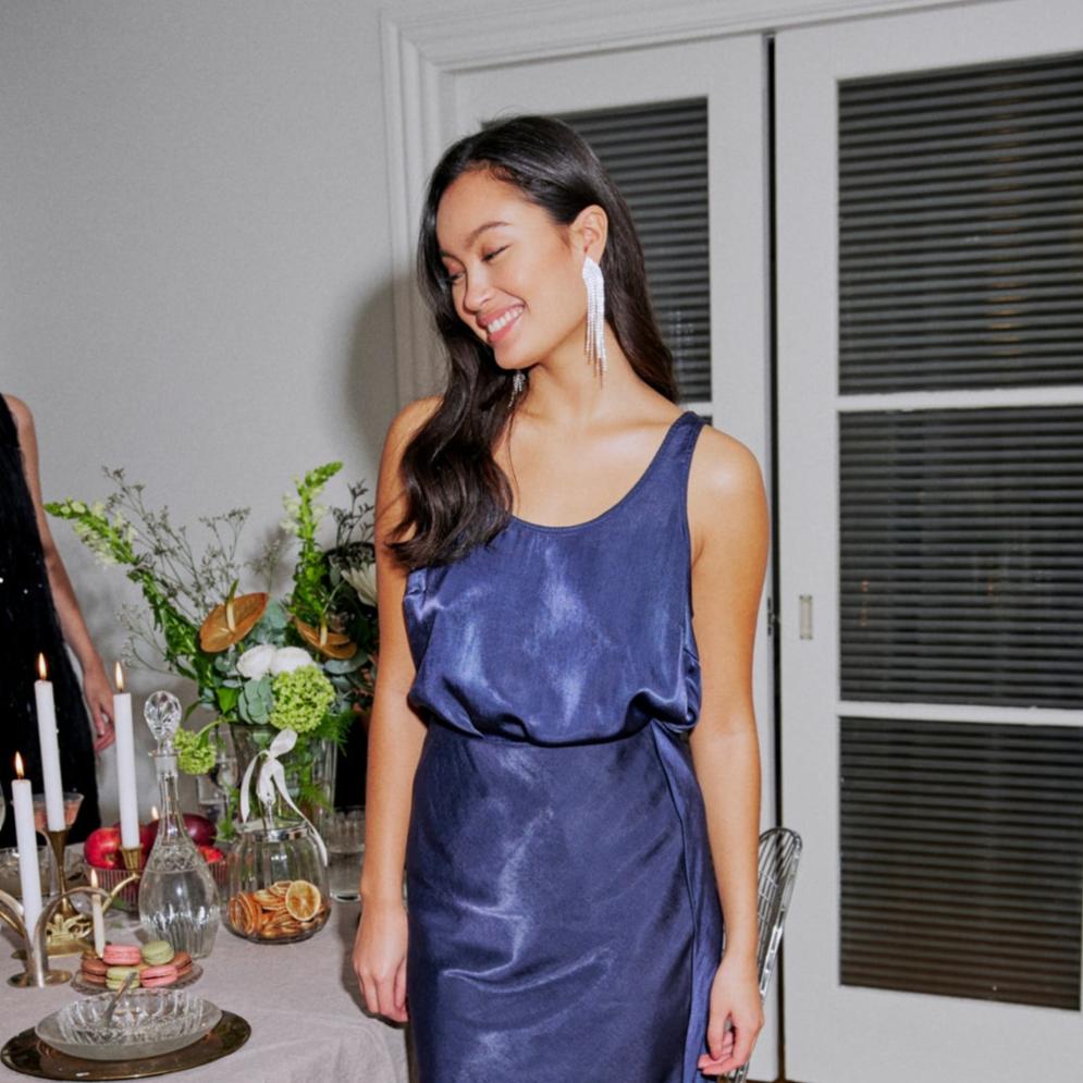 A woman smiles in a sleeveless navy blue Fransa dress, surrounded by candles and floral arrangements.