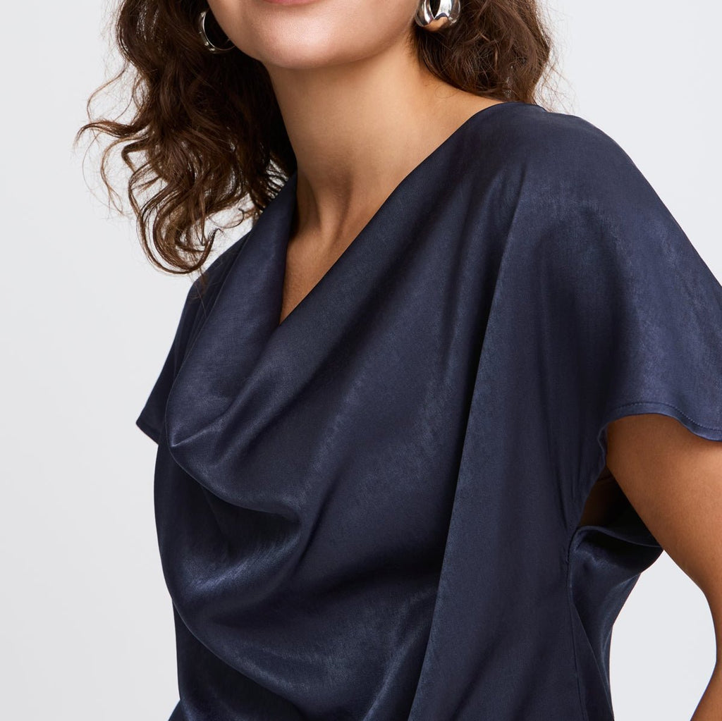 A woman wears a navy blue satin top by Fransa, featuring a draped neckline and short sleeves.