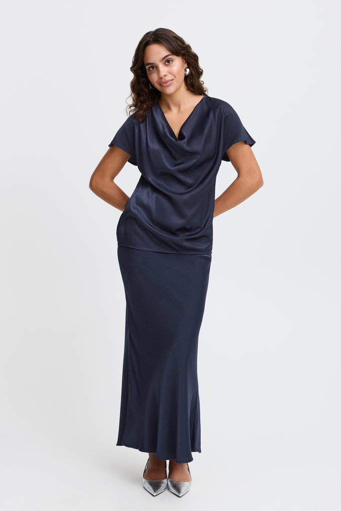 A woman stands in a navy dress by Fransa with a cowl neckline and silver heels, smiling at the camera.