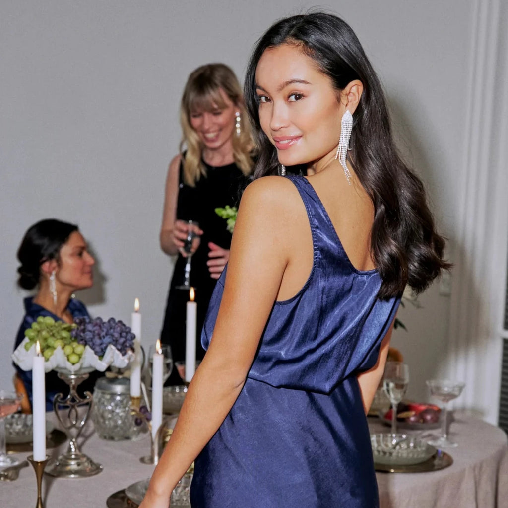 A woman in a navy Fransa dress smiles at the camera, with guests in black attire at a dinner table.