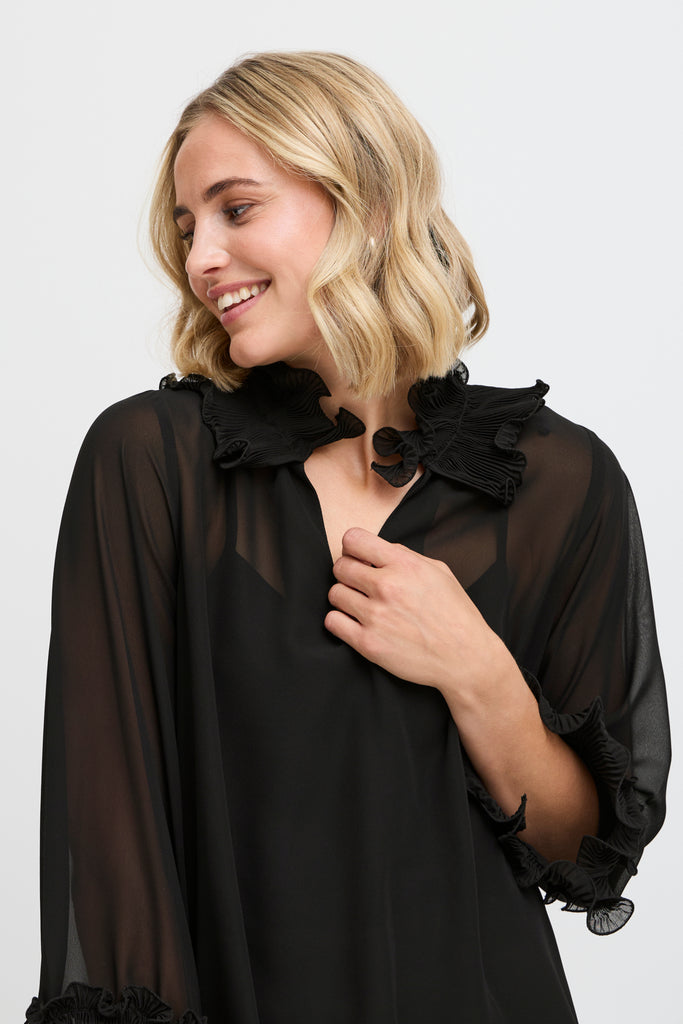 A smiling woman wearing a black Fransa blouse with ruffled details at the collar and sleeves.