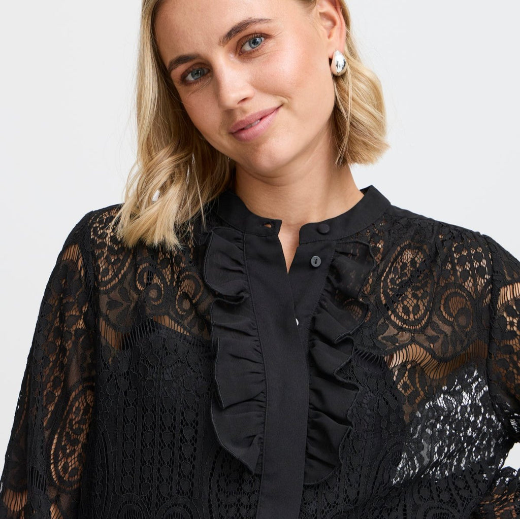 A woman wearing a black Fransa blouse with lace details and ruffles, smiling softly.
