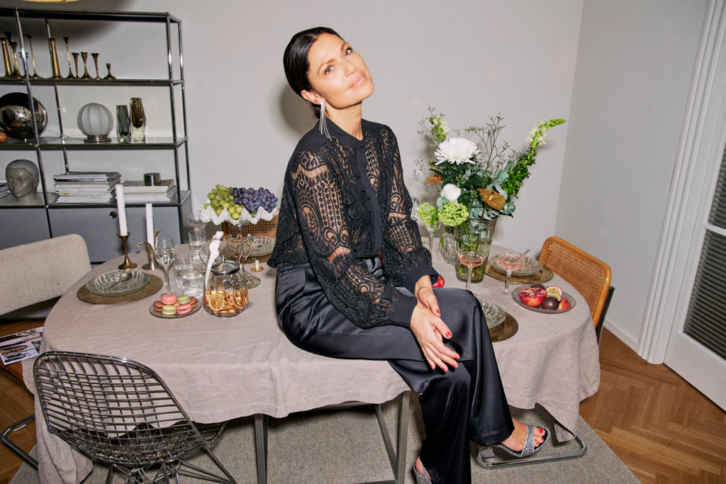 Woman sits at a table wearing a black Fransa lace top and black satin pants, surrounded by flowers and fruit.
