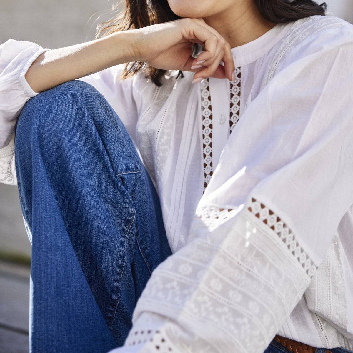 Woman wearing a white blouse with lace details and blue jeans, sitting outdoors.